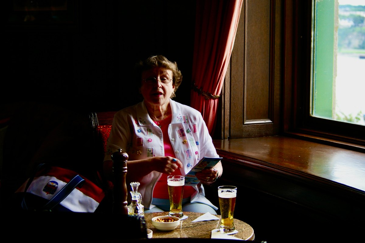 Carol beside a window with her bag and two beers on the table in front of her