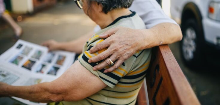 A woman sitting on a bench and looking at pictures, with a loved one's arm around her shoulder