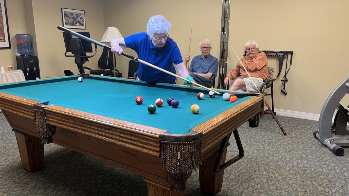 A woman attempting a tricky billiards shot while two men watch, in the pool room at Branchlands' Manor House.