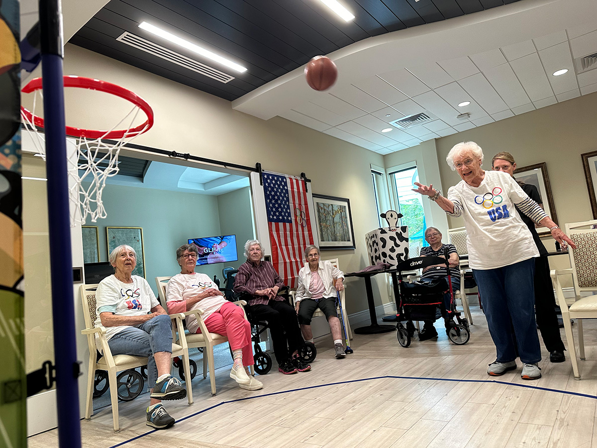 An assisted living resident throwing a small basketball into a hoop while friends watch.